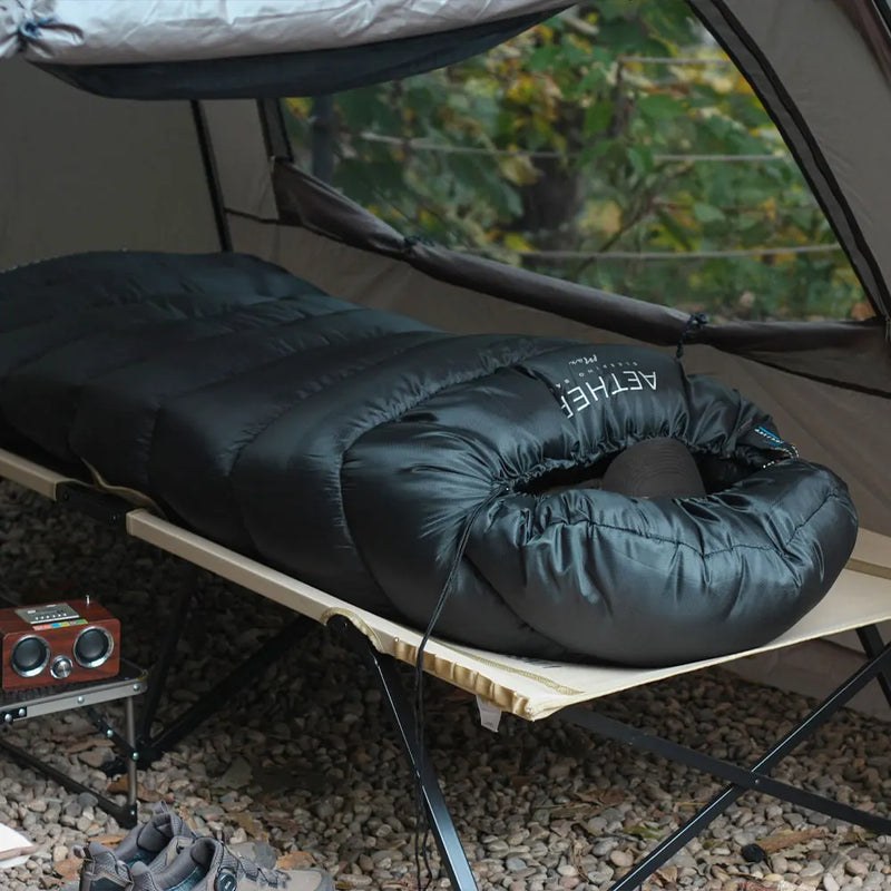 Load image into Gallery viewer, Black sleeping bag with a neck pillow on a camping cot outdoors.
