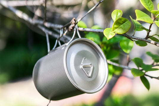 Metallic camping mug with a lid hanging on a tree branch with green leaves.