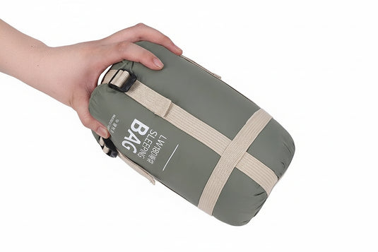 Hand holding a green sleeping bag with beige straps on a white background