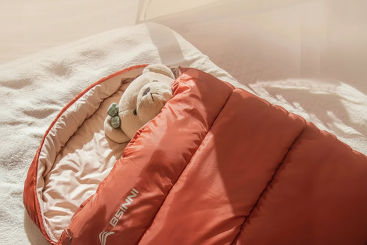 Person lying in a large red sleeping bag with a teddy bear, labeled '100 cm', on a soft surface.