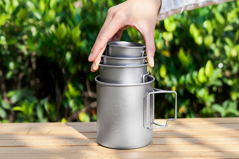 Load image into Gallery viewer, Stackable titanium mug with handle held by a hand on a wooden surface with greenery in the background
